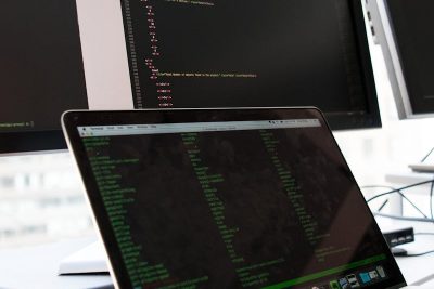 A developer writes code on a laptop in front of multiple monitors in an office setting.